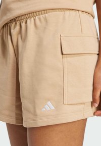 Béžové bavlněné šortky s elastickým pasem, boční kapsou a bílým logem Adidas na lemu. Hladká textura a volný design.