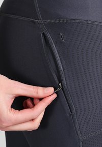 Mustat urheilulegginsit, joissa on vetoketjullinen sivutasku, teksturoitu paneeli geometrisella kuviolla ja sileä kankaan koostumus.