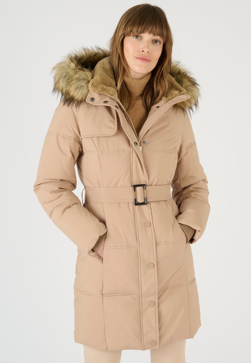 Manteau rembourré beige avec une capuche en fourrure, taille ceinturée, fermeture éclair et accents de boutons, présentant des sections matelassées et des poches latérales.