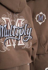 Brun sweatshirt med stor broderet "Multiply" tekst i blå og creme, med en udsmykket "M" på ryggen. Blødt stof og afslappet pasform.