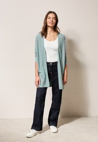 Ljusblå cardigan med långa ärmar och ficka fram, tillsammans med högmidjade mörka jeans och vita sneakers.