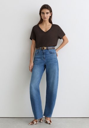 Vrouw staat tegen een witte muur, draagt een bruin V-hals T-shirt, blauwe high-waisted jeans met riem en hakken met koeienprint.