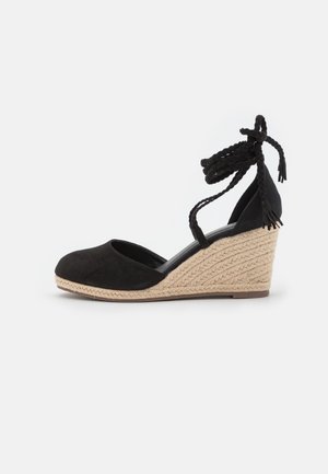 Chaussure à talon compensé en daim noir avec bout arrondi, plateforme enveloppée de jute et liens tressés à la cheville. Texture lisse avec design minimaliste.