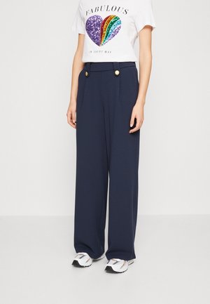 Wit T-shirt met kleurrijk sequentien hart en tekst "Fabulous", marineblauwe wijd uitlopende broek met gouden knopen en witte sneakers gedragen door een staand persoon.