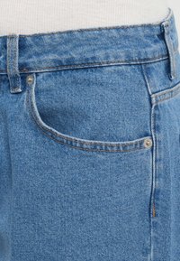 Sinine denimist teksad klassikalise lõikega, mis sisaldavad esikotti, hõbedast riistvara ja nähtavat õmblust. Tekstuur on pehme ja sile.