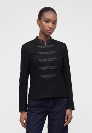 Jeune femme portant une veste style militaire noire boutonnée avec des garnitures en satin et un pantalon bleu foncé, debout devant un fond uni.