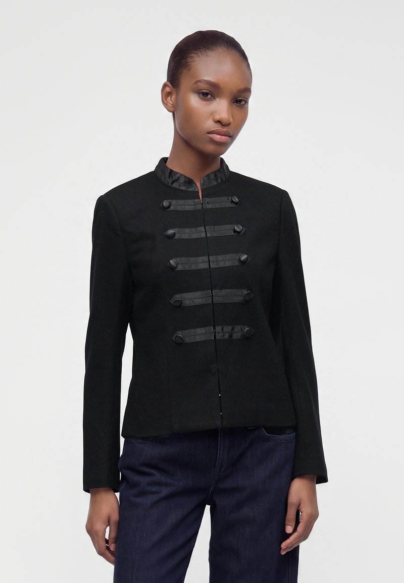 Jeune femme portant une veste style militaire noire boutonnée avec des garnitures en satin et un pantalon bleu foncé, debout devant un fond uni.