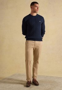Homme debout, les mains dans le dos, portant un pull torsadé bleu marine, un pantalon beige et des mocassins marron sur un fond neutre.