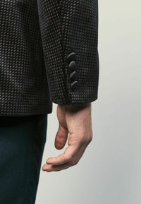 Mustan tekstuurin bleiserin hiha, jossa on hillitty geometrinen kuvio, neljällä pyöreällä kankaasta päällystetyllä napilla ja muotoillulla istuvuudella.