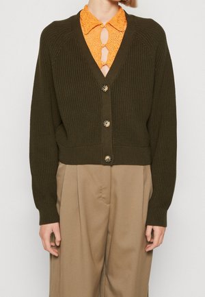 Person trägt eine dunkelolivgrüne, gestrickte Strickjacke mit Knöpfen über einem orangen Spitzenoberteil und beigen, hoch taillierten, plissierten Hosen.