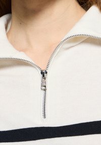 Gros plan sur un haut en maille blanche partiellement zippé avec une bande horizontale noire traversant la poitrine.