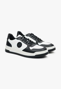 Černobílé kožené tenisky s perforovanou špičkou, černými tkaničkami a kulatým logem na boku. Silná gumová podrážka pro lepší trakci.