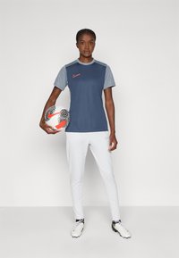 Nike urheiluasu, jossa on laivastonsininen ja vaaleanharmaa lyhythihainen paita, jossa on oranssi logo, yhdistettynä valkoisiin housuihin ja mustiin nappulakenkiin.