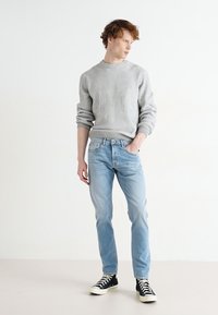 Pull en maille côtelée gris clair, jeans bleu clair et baskets montantes noires. La tenue présente une coupe décontractée avec une texture et des détails subtils.