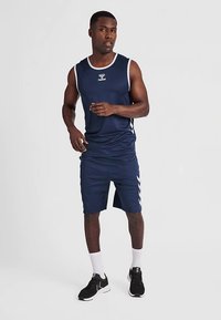 Débardeur de sport bleu marine avec accents blancs, assorti à un short avec motifs chevrons sur les côtés. Le modèle porte des baskets noires et des chaussettes blanches.