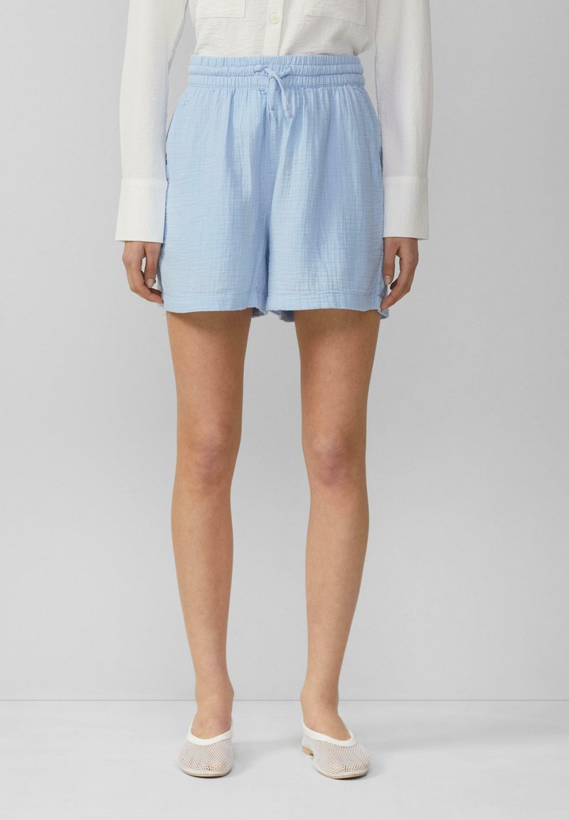 Shorts en coton bleu clair avec une taille élastique et un cordon de serrage. Présentent une finition texturée et des poches latérales. Associés à des chaussures slip-on en maille blanches.