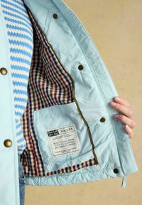 Mano sosteniendo una chaqueta azul claro abierta que muestra el forro interior a cuadros y una etiqueta blanca con la marca y texto dentro del bolsillo.