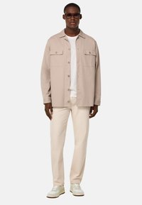 Camicia beige con bottoni e tasche sul petto, abbinata a pantaloni crema chiaro e sneakers bianche. Vestibilità casual e tessuto dalla superficie liscia.