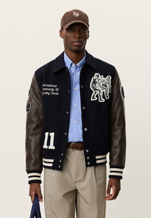 Marineblå baseballjakke i uld med brune læderærmer, hvid tekst og et grafisk design af to figurer. Kombineret med en blåstribet skjorte.