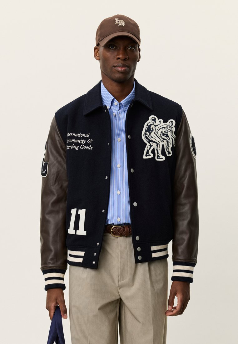 Μπλε ναυτικό μάλλινο μπέιζμπολ μπουφάν με καφέ δερμάτινα μανίκια, λευκό κείμενο και γραφιστικό σχέδιο δύο μορφών. Συνδυασμένο με μπλε ριγέ πουκάμισο.