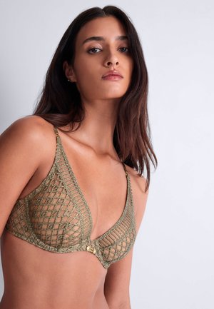 Jeune femme aux longs cheveux foncés portant un soutien-gorge vert à motif en maille, regardant avec confiance vers la caméra sur un fond uni.