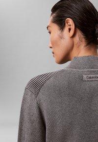 Szary sweter o prążkowanej fakturze z paskami na ramionach. Posiada wysoki kołnierz oraz metkę z napisem "Calvin Klein" z tyłu.
