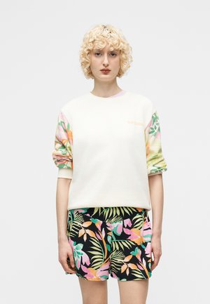 Άτομο με ξανθά, σγουρά μαλλιά, που φοράει λευκό φούτερ με ανθισμένα μανίκια και μαύρο σορτς με πολύχρωμα τροπικά φύλλα, στέκεται όρθιο κοιτώντας μπροστά.