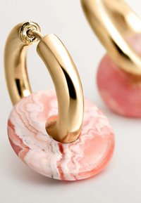 Pendientes de aro dorados que presentan una piedra circular de mármol rosa, mostrando una textura suave y sutiles variaciones en color y patrón.