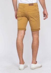 Shorts en coton beige avec des ourlets retroussés et des poches arrière, présentant des détails de couture subtils. Portés avec des baskets blanches sur un fond neutre.
