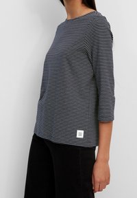Frau in einem navy- und weiß gestreiften, dreiviertelärmeligen Oberteil und schwarzen Hosen, seitliche Ansicht mit sichtbarem Bekleidungslabel.