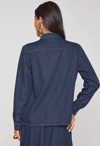 Giacca in denim scuro con colletto classico, maniche lunghe e polsini con bottoni. La vista posteriore mostra le cuciture e una vestibilità rilassata.