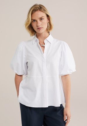 Donna bionda che indossa una camicetta bianca con maniche a sbuffo e colletto e pantaloni neri, accessoriata con orecchini a cerchio d'argento e collana.