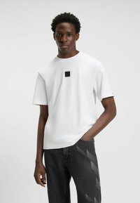 Weißes Baumwoll-T-Shirt mit kurzen Ärmeln, einem Rundhalsausschnitt und einem kleinen schwarzen Logo-Detail auf der Brust. Kombiniert mit dunkelgrauen Jeans.