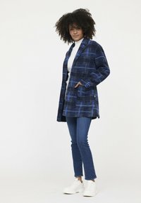 Blauw geruit jasje met een ontspannen pasvorm, twee voorzakken en een reverskraag, gedragen over een witte trui, gecombineerd met blauwe jeans en witte sneakers.