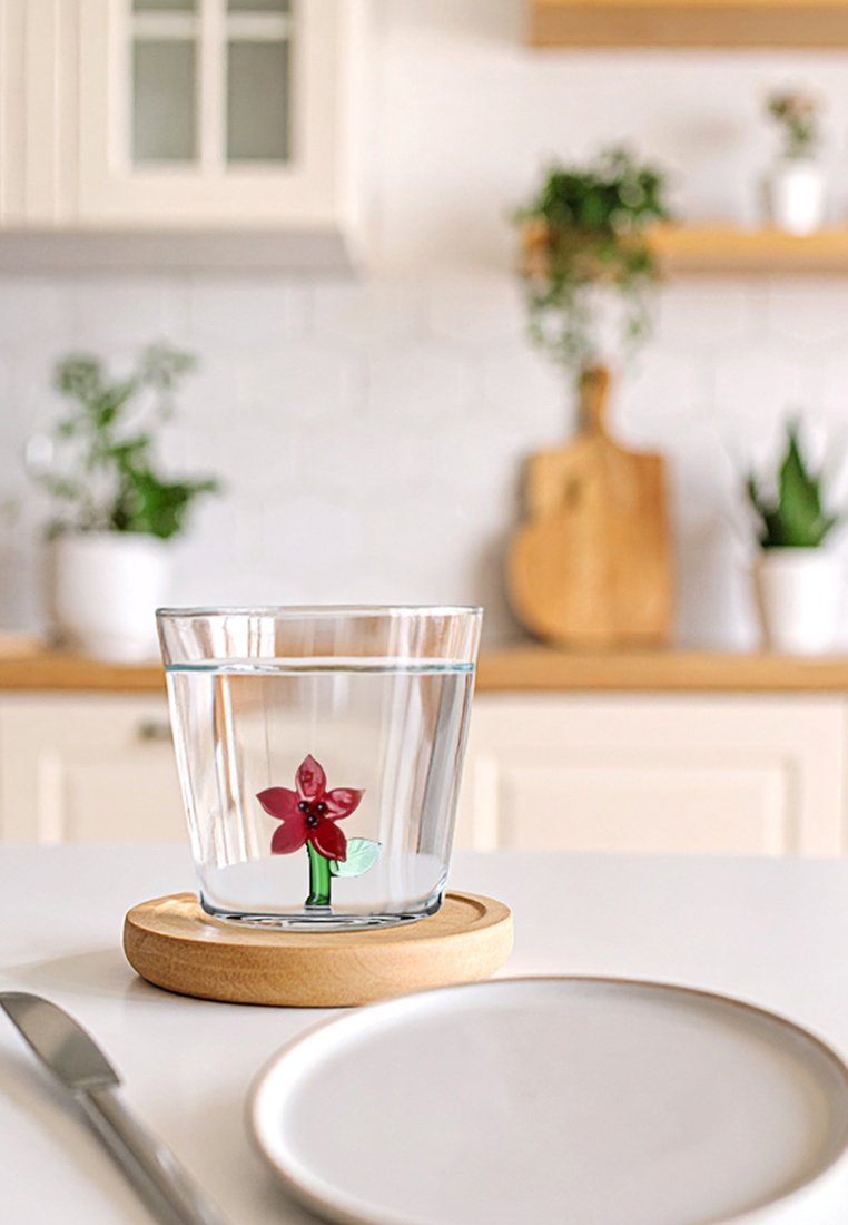 Przezroczysta szklanka na okrągłym drewnianym podstawku, wypełniona wodą i ozdobiona czerwonym wzorem kwiatu z zielonymi liśćmi w środku. Obok biały talerz i nóż.