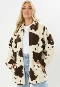 Nepbont jas in crème met een bruin koeienpatroon, voorzien van een kraag, lange mouwen en zijzakken, gecombineerd met hoge taille jeans.