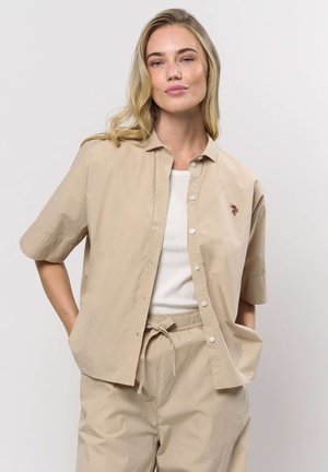 Femeie blondă purtând o cămașă bej cu mâneci scurte și nasturi, și pantaloni asortați cu șnur, pozând cu mâinile în buzunare, pe un fundal alb.