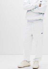 Lichtgrijze sweatshirt en bijpassende joggingbroek van zacht materiaal, met een klein logo op de borst en aan de zijkant, gecombineerd met witte sneakers.