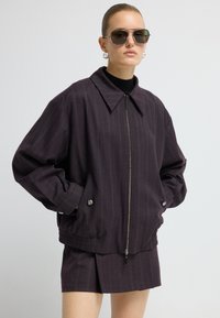 Paarse geblokte bomberjack met een brede kraag, zilveren rits en zijzakken; gedragen over een zwarte coltrui en een bijpassende rok.