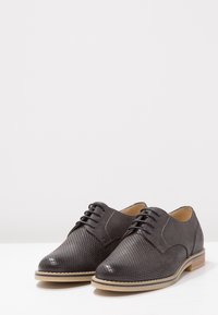 Scarpe in pelle grigio scuro stringate con design superiore perforato, punta arrotondata e suola beige chiaro con accentuazioni di cuciture in contrasto.