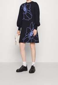 Abito nero con maniche oversize, caratterizzato da contorni floreali azzurri. Abbinato a scarpe nere e calzini bianchi, mentre tiene una borsa con frange.