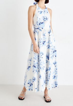 Femme portant une robe longue blanche à col halter avec imprimé floral bleu et des sandales argentées plates à brides, debout devant un fond blanc uni.