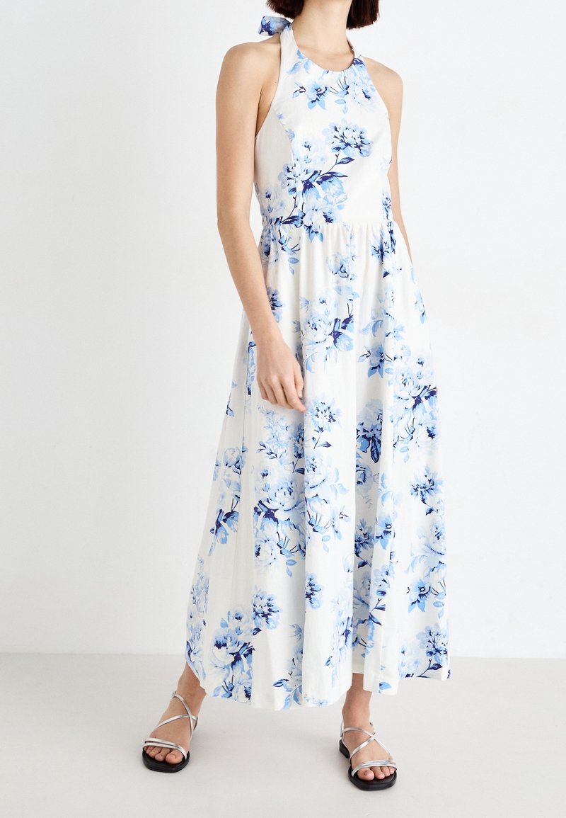 Femme portant une robe longue blanche à col halter avec imprimé floral bleu et des sandales argentées plates à brides, debout devant un fond blanc uni.