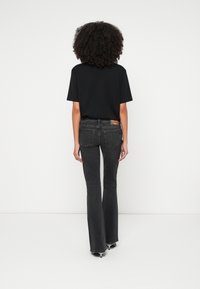 Personne aux cheveux bouclés de dos, portant un t-shirt noir, un jean foncé évasé et des chaussures à talons sur fond blanc uni.