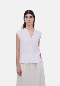 Top de lino blanco con mangas cortas y escote en forma de V. Presenta bordes de mangas en forma de ondas y un corte relajado. Textura suave. Color claro.