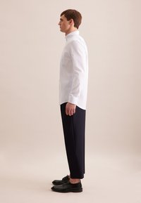 Wit en lichtblauw gestreept overhemd met lange mouwen, gecombineerd met marineblauwe broek en zwarte veterschoenen, gezien van opzij.