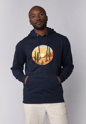 Mann trägt marineblauen Kapuzenpullover mit Grafik eines Wüstensonnenuntergangs und Kaktus, Hände in der Fronttasche, beige Hose, neutraler grauer Hintergrund.