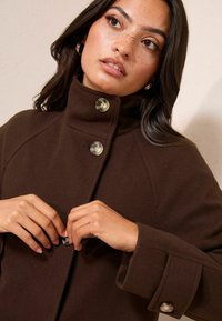 Rudas vilnos mišinio paltas su aukštu apykakliu ir dviem dideliais mygtukais, pasižymintis laisvu kirpimu ir struktūruotomis rankovėmis. Minkšta tekstūra ir apdaila.