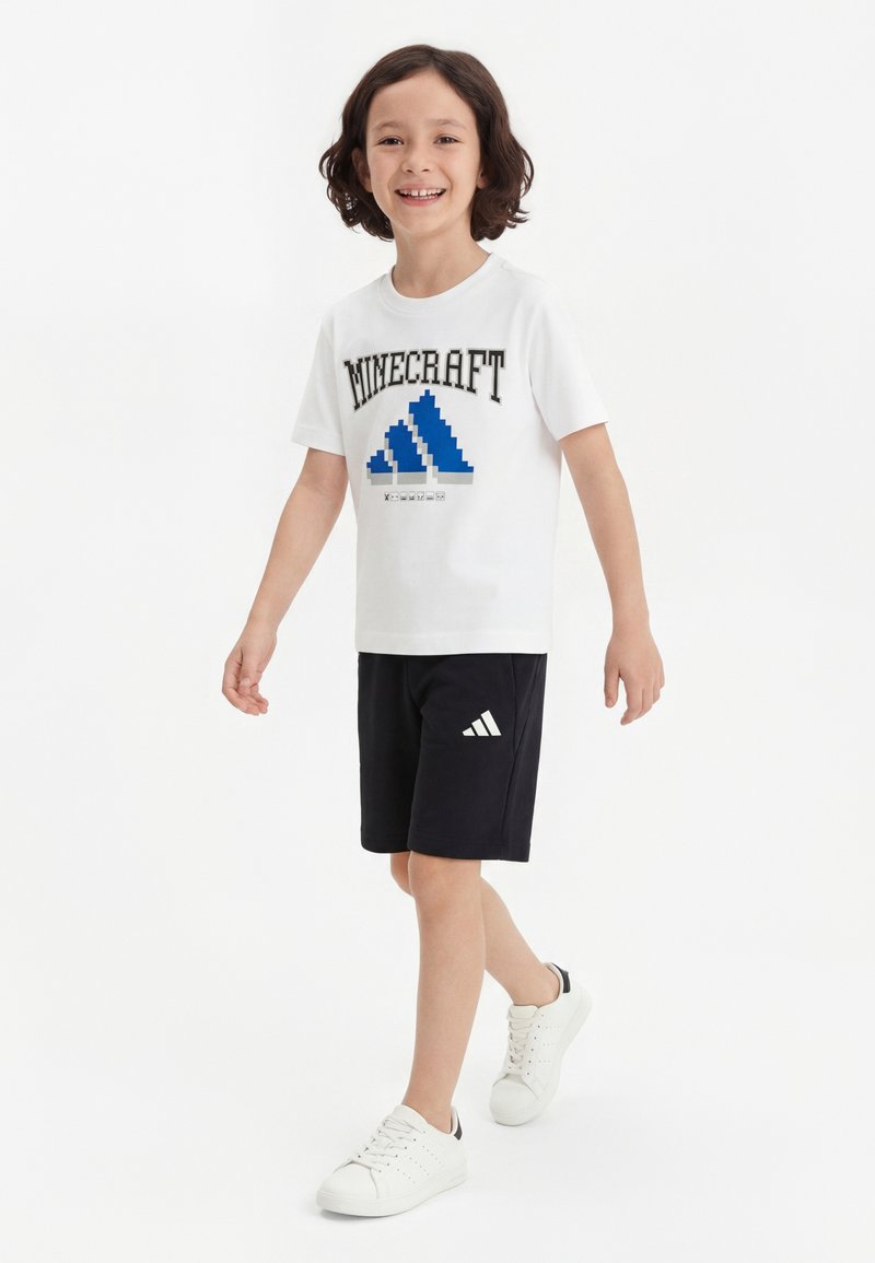 Garçon marchant et souriant, portant un t-shirt blanc Minecraft, un short noir Adidas et des baskets blanches sur fond blanc uni.