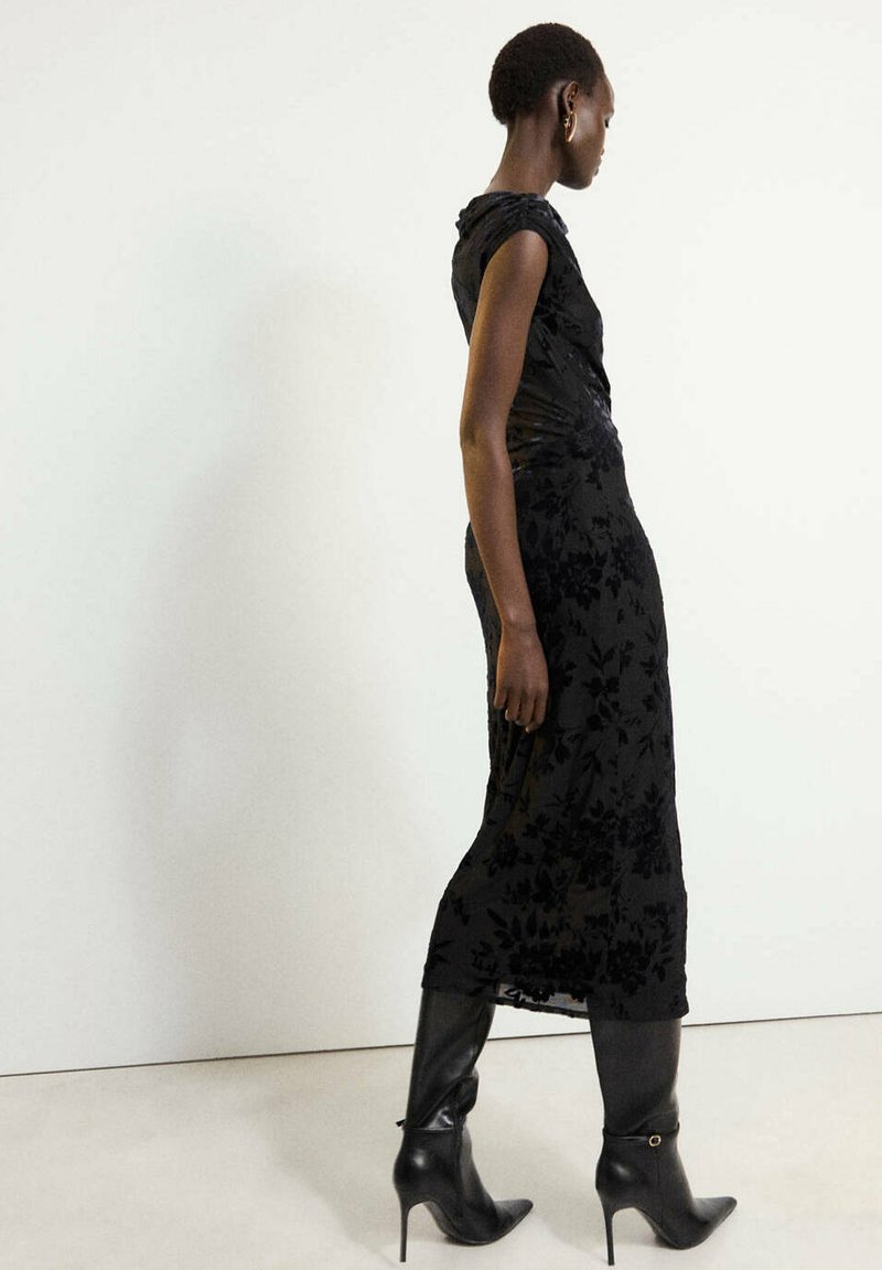 Un mannequin marche de profil, vêtu d’une robe midi noire à motifs et de bottes à talons montant jusqu’aux genoux, devant un fond de studio uni.